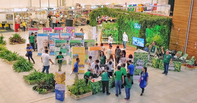 Personas caminando por una exposición en un salón de eventos con plantas y casetas de la feria del libro.