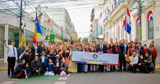 A large group pf people, some holding flags and a banner, pose for a photo in a street.