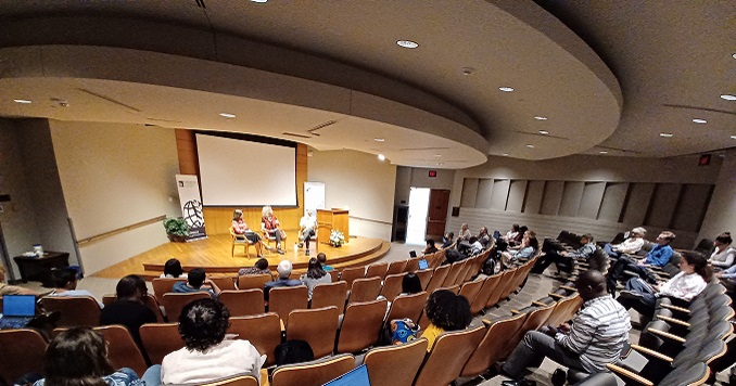 People sitting in a conference room and a panel of three speakers on a stage.