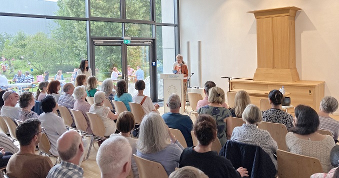 A few dozen people seated, listening to a person speaking at a podium in a room with a Buddhist altar.