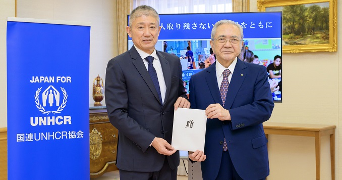 Dos hombres con un sobre en la mano junto a una pancarta de la agencia japonesa del ACNUR.