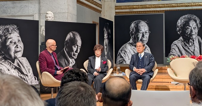 Three people seated on a stage with large photographs in the background showcasing hibakusha portraits.