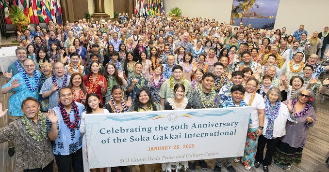 People standing in an auditorium, smiling and wearing traditional floral garlands.