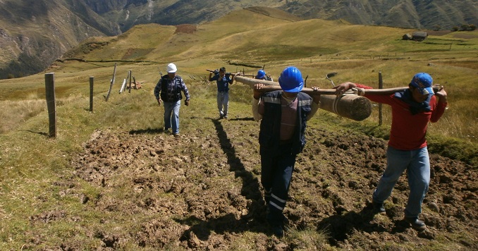 Men carrying a wooden pole uphill through a field surrounded by a mountainous landscape.