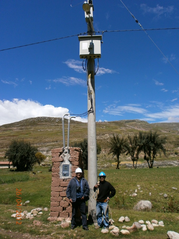 Two men in hard hats standing on opposite sides of a transmission pole in the countryside, with clear skies and expansive fields in the background.