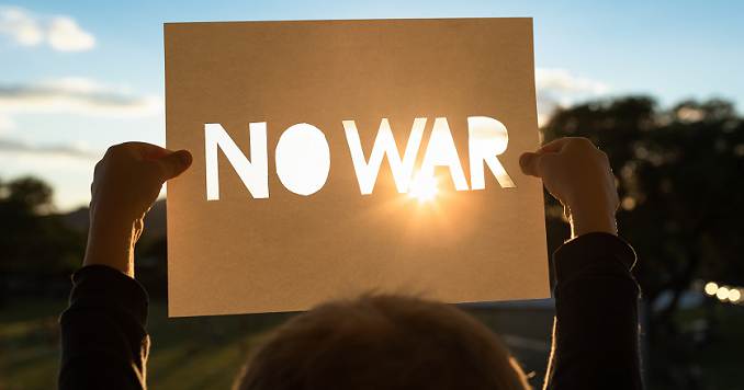 A person holding up a paper cut-out reading “No War,” silhouetted against the bright sun.