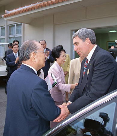 Two men shaking hands to greet each other in front of a building and some people in the background.