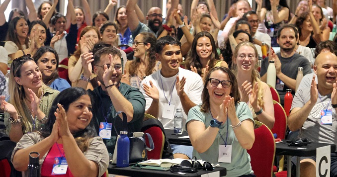 Muchos jóvenes en una sala de conferencias, levantan los brazos y aplauden.