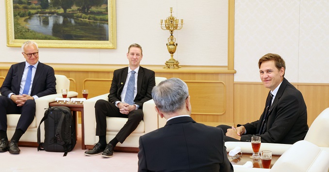 Cuatro hombres con trajes conversan en una gran sala con sillones blancos.