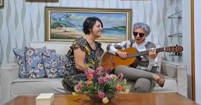 Una mujer con el pelo gris y gafas oscuras toca la guitarra para su amiga.