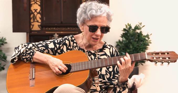 Una mujer con gafas oscuras toca la guitarra sentada.