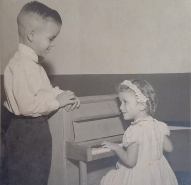 Fotografía en blanco y negro de un niño mirando a su hermana que está tocando un pequeño piano.