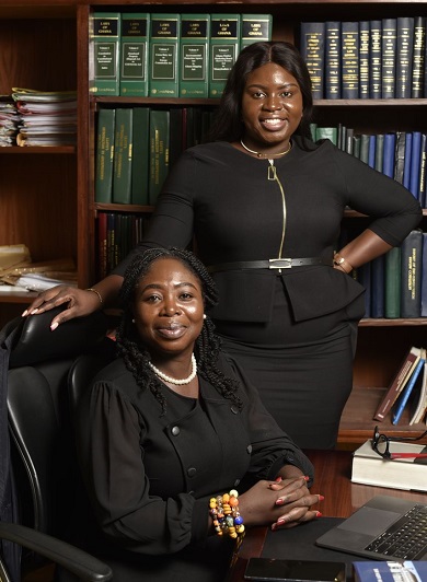 Two women dressed in formal clothes in an office in front of some book-filled shelves.