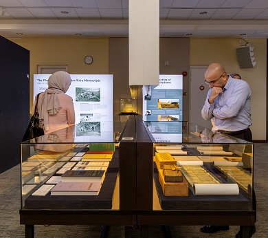 In a dimly lit room, one person viewing scrolls in a glass case and another viewing an exhibition panel.