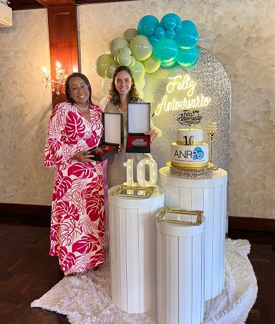 Two formally dressed women stand smiling together in front of a cake at a celebration.