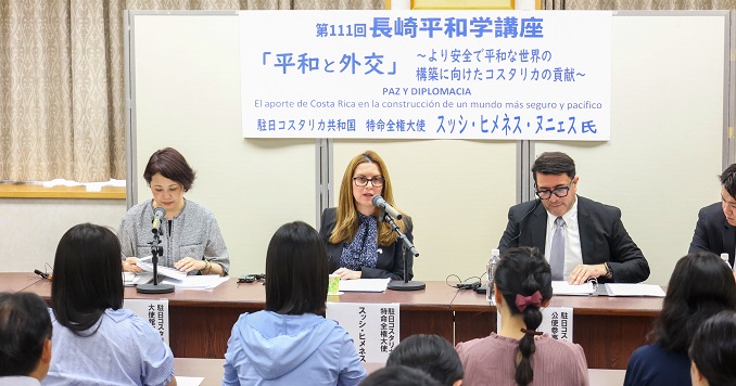 一名女性坐在桌前与其他专题发言人一同面向现场听众发表演说。