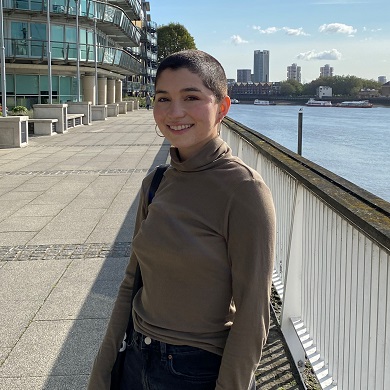 Isabella stands on a paved pathway beside a river with city buildings in the background.