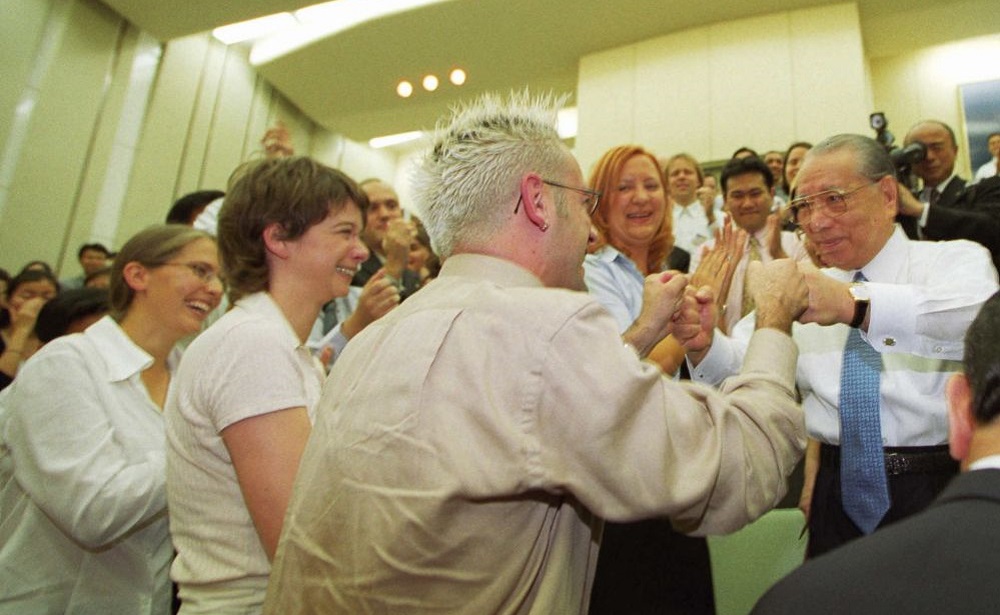 Mr. Ikeda stands among smiling youth members in an auditorium, fist-bumping with a young man