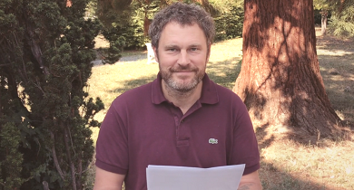 A man seated beneath a tree reads to the camera