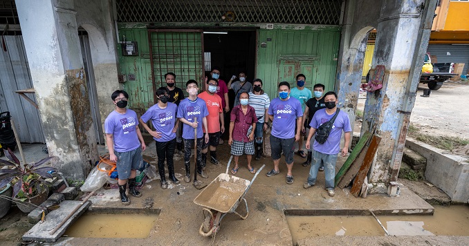 A group photo of a dozen men and young men with a wheelbarrow outside a flood-damaged house.